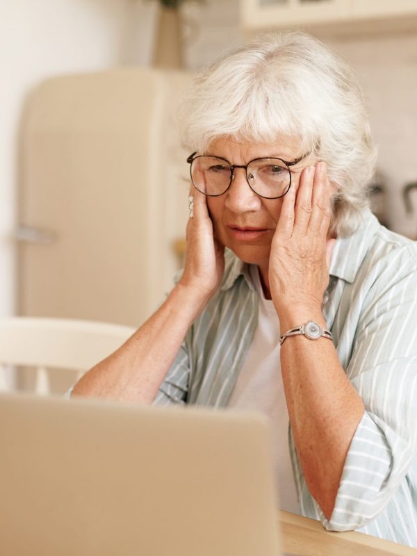 Lonely upset retired woman with gray hair having frustrated stressed look, touching head, facing financial problems, trying to save money to pay off all her debts, calculating expenses, using laptop